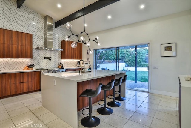 kitchen with wall chimney exhaust hood, decorative light fixtures, a kitchen bar, an island with sink, and beam ceiling