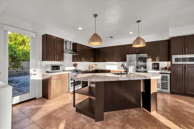 kitchen featuring an island with sink, beverage cooler, hanging light fixtures, stainless steel appliances, and wall chimney exhaust hood