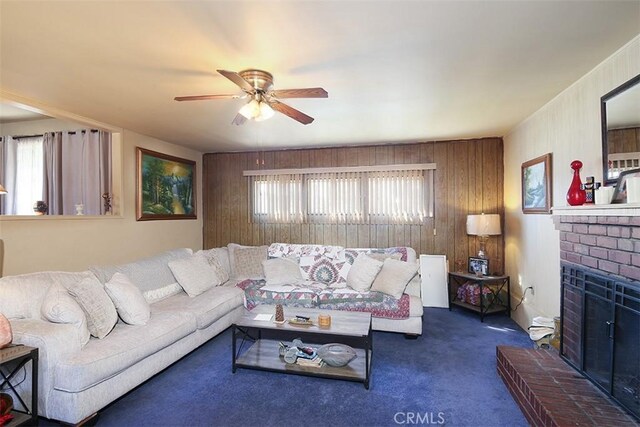 living room featuring ceiling fan, a fireplace, dark carpet, and wooden walls