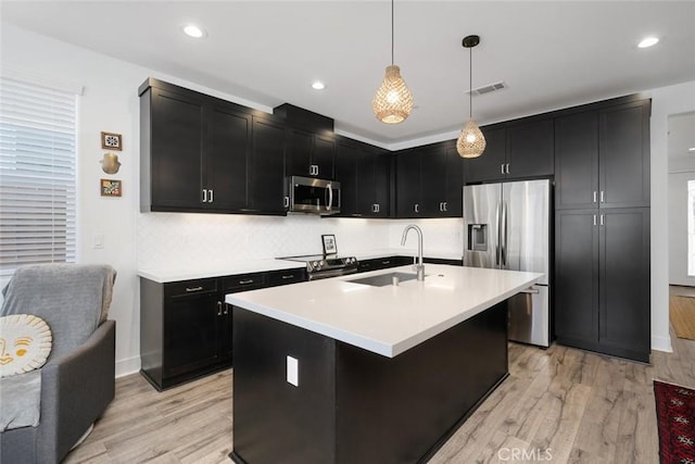 kitchen with appliances with stainless steel finishes, decorative light fixtures, an island with sink, light hardwood / wood-style floors, and sink