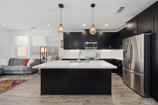 kitchen featuring appliances with stainless steel finishes, an island with sink, sink, hanging light fixtures, and light hardwood / wood-style flooring