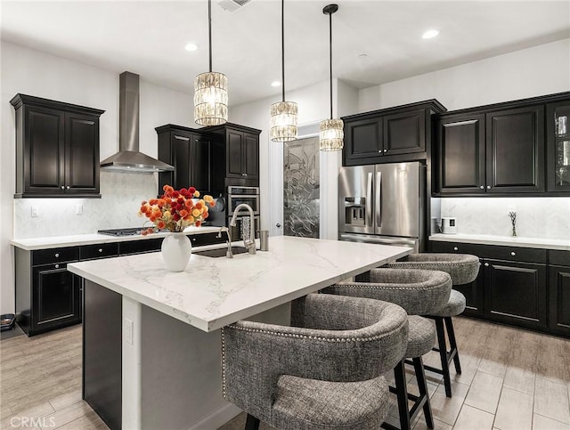 kitchen with backsplash, wall chimney range hood, sink, appliances with stainless steel finishes, and an island with sink