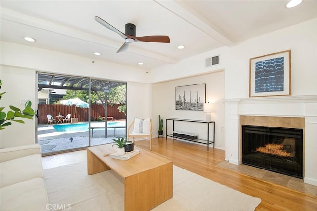 living room with ceiling fan, a tile fireplace, beam ceiling, and light hardwood / wood-style flooring