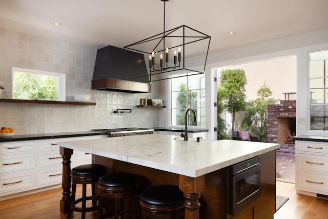 kitchen featuring decorative backsplash, premium range hood, white cabinetry, and an island with sink