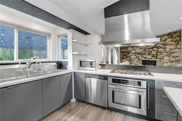 kitchen with wall chimney range hood, gray cabinetry, light wood-style floors, decorative backsplash, and stainless steel appliances