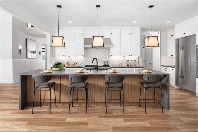 kitchen featuring white cabinetry, high end fridge, a kitchen island with sink, wall chimney range hood, and sink