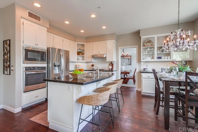 kitchen featuring visible vents, white cabinets, glass insert cabinets, a kitchen island with sink, and stainless steel appliances