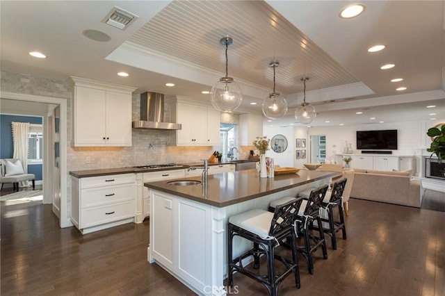 kitchen with a raised ceiling, white cabinets, wall chimney range hood, and a center island with sink