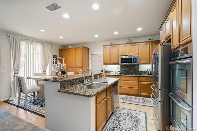 kitchen featuring light tile patterned floors, stainless steel appliances, a kitchen island with sink, dark stone countertops, and sink