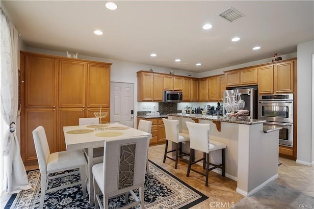 kitchen with a kitchen bar, a center island with sink, appliances with stainless steel finishes, decorative backsplash, and light stone counters