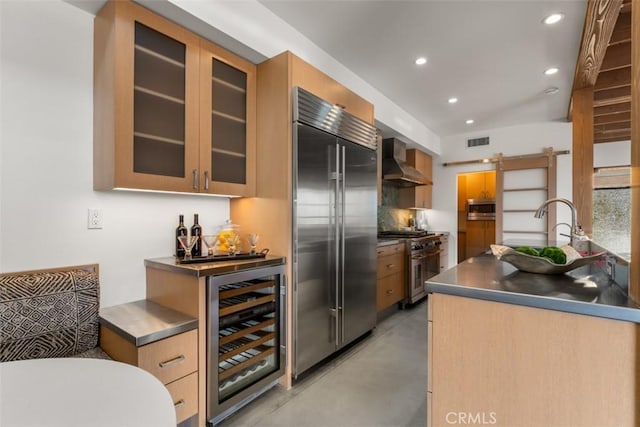 kitchen featuring stainless steel counters, wall chimney range hood, built in appliances, wine cooler, and a barn door