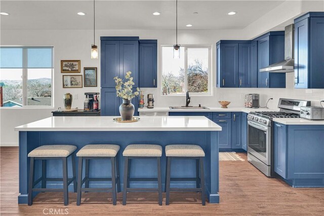 kitchen with wall chimney range hood, a kitchen bar, blue cabinets, stainless steel gas range oven, and a sink