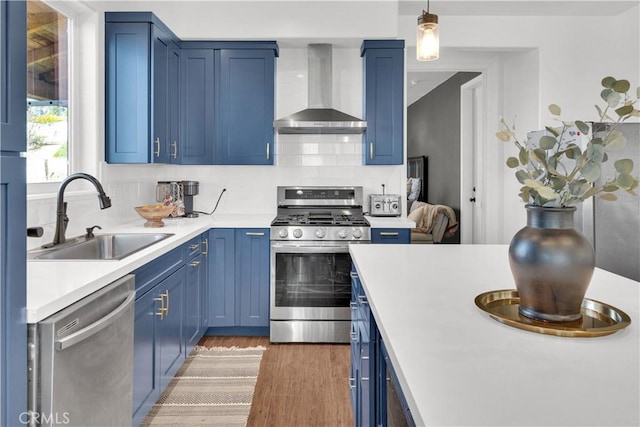 kitchen with blue cabinets, stainless steel appliances, a sink, wall chimney exhaust hood, and light wood finished floors