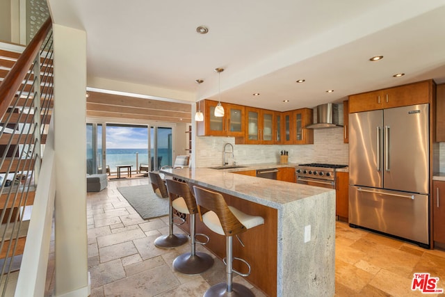 kitchen featuring wall chimney exhaust hood, high end appliances, hanging light fixtures, kitchen peninsula, and light stone counters