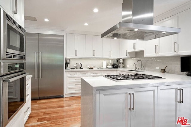 kitchen with island exhaust hood, backsplash, built in appliances, white cabinets, and sink