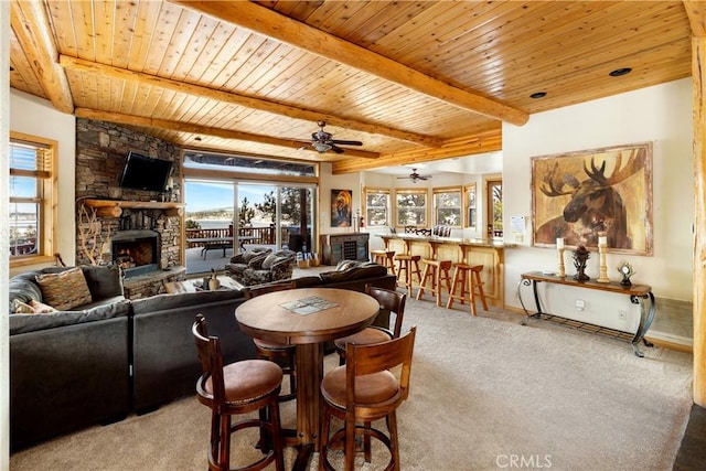 carpeted dining room featuring ceiling fan, wood ceiling, beam ceiling, and a stone fireplace