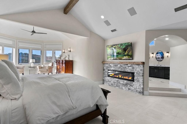 bedroom with ceiling fan, beam ceiling, a stone fireplace, connected bathroom, and high vaulted ceiling