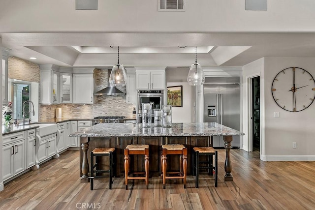 kitchen featuring appliances with stainless steel finishes, wall chimney exhaust hood, a kitchen island, sink, and a raised ceiling