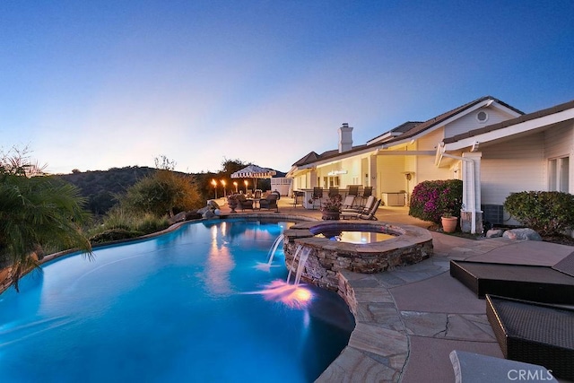 pool at dusk featuring pool water feature, a patio area, and an in ground hot tub