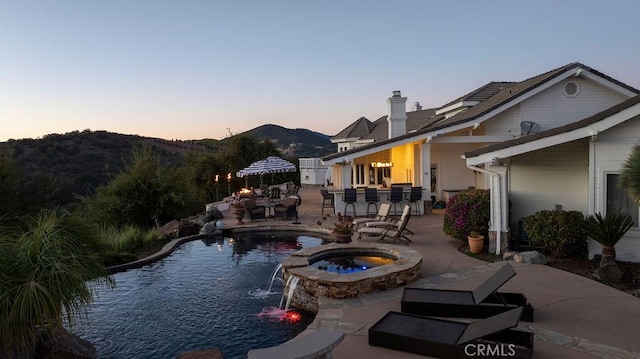 pool at dusk featuring a patio area, an in ground hot tub, pool water feature, a mountain view, and a bar