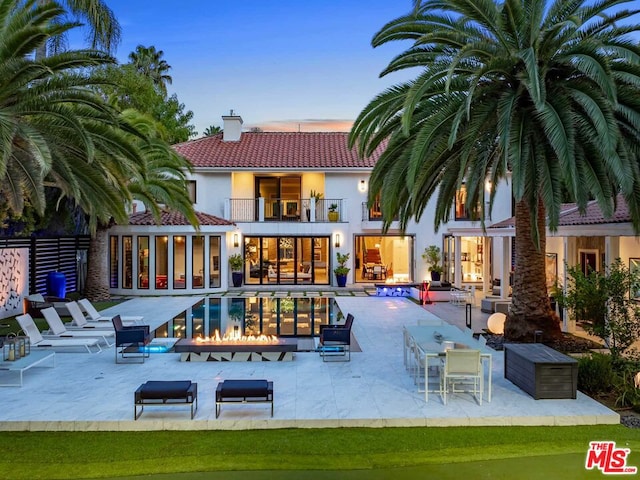 back house at dusk with a balcony, a patio area, an outdoor fire pit, and a swimming pool