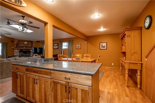 kitchen with light hardwood / wood-style floors, a stone fireplace, ceiling fan, dark stone counters, and sink