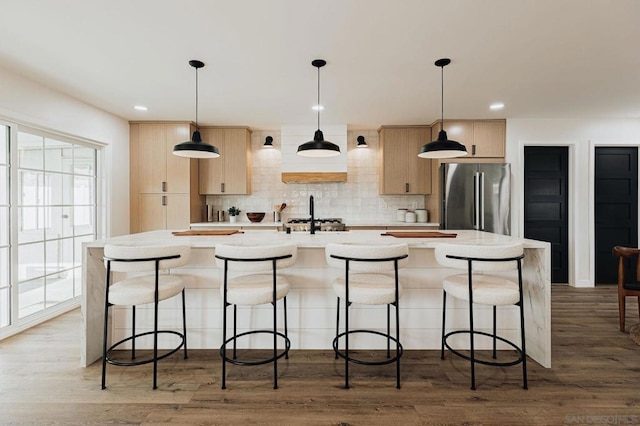 kitchen featuring light brown cabinetry, hanging light fixtures, a kitchen island with sink, stainless steel refrigerator, and hardwood / wood-style flooring