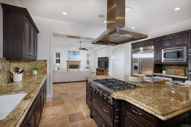 kitchen featuring stone tile floors, open floor plan, built in appliances, light stone countertops, and island exhaust hood