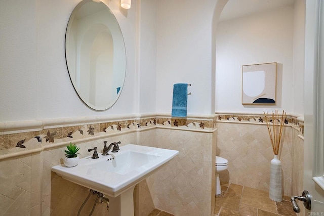 bathroom with tile walls, toilet, stone finish flooring, wainscoting, and a sink