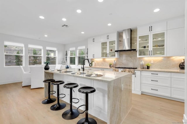 kitchen featuring light stone countertops, wall chimney range hood, an island with sink, light hardwood / wood-style floors, and decorative backsplash