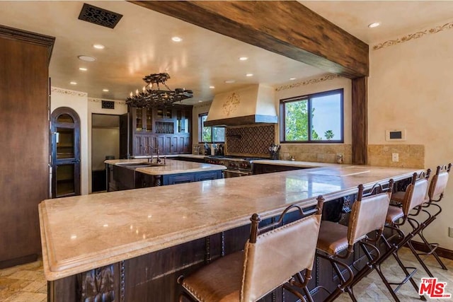 kitchen featuring backsplash, kitchen peninsula, a kitchen breakfast bar, an island with sink, and custom range hood