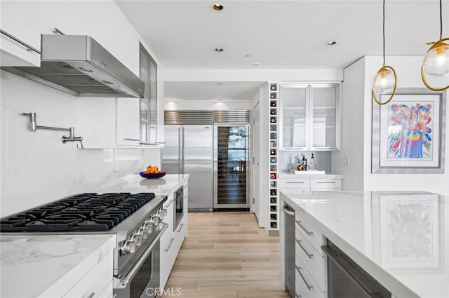 kitchen featuring premium appliances, light stone countertops, pendant lighting, white cabinets, and wall chimney range hood