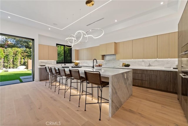 kitchen with light hardwood / wood-style flooring, a kitchen bar, backsplash, a tray ceiling, and a center island with sink