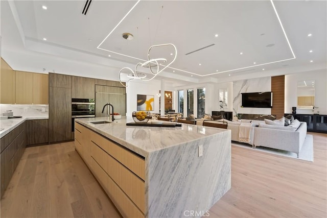 kitchen with a large island with sink, a raised ceiling, light hardwood / wood-style flooring, light stone counters, and black oven