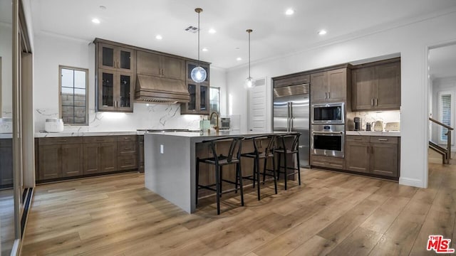 kitchen with custom exhaust hood, light hardwood / wood-style floors, an island with sink, decorative light fixtures, and built in appliances