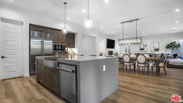 kitchen with a center island with sink, light wood-type flooring, hanging light fixtures, built in appliances, and dark brown cabinetry