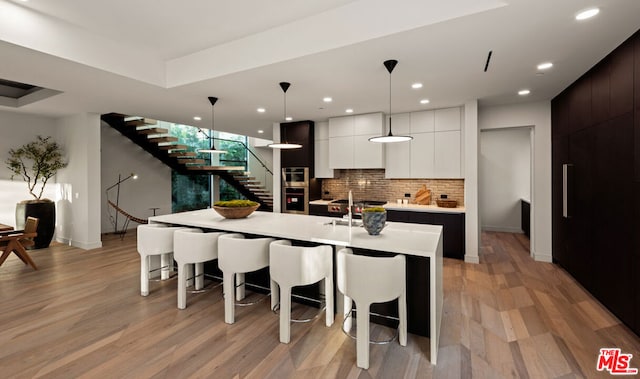 kitchen featuring light hardwood / wood-style floors, an island with sink, dark brown cabinets, pendant lighting, and white cabinets