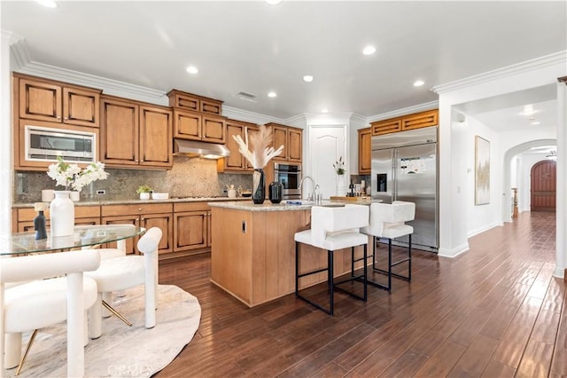 kitchen with a kitchen bar, a center island with sink, crown molding, and built in appliances