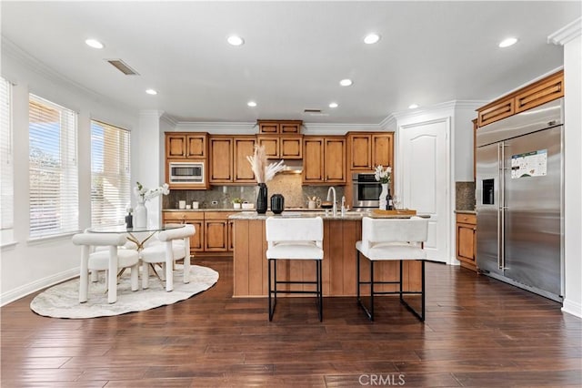 kitchen with crown molding, a wealth of natural light, built in appliances, and a center island with sink