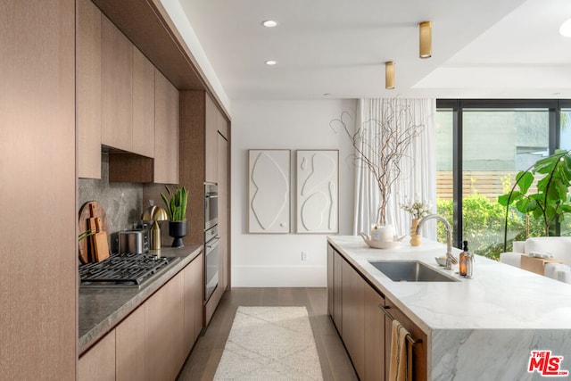 kitchen with backsplash, stainless steel gas stovetop, sink, a kitchen island with sink, and light stone counters