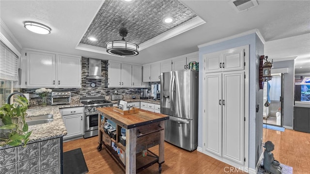 kitchen with crown molding, a tray ceiling, appliances with stainless steel finishes, white cabinets, and wall chimney exhaust hood