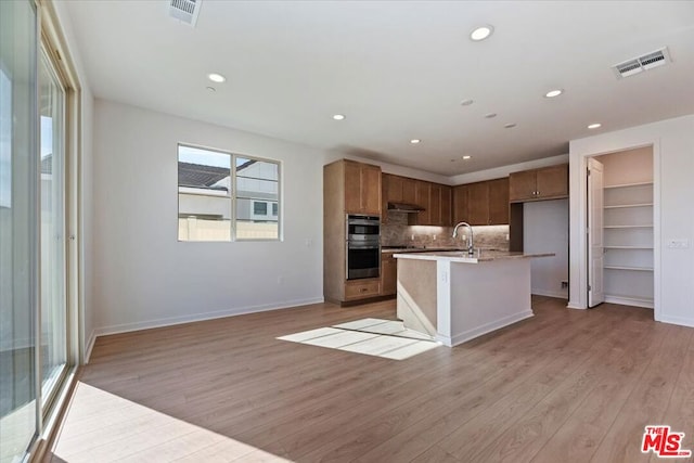 kitchen with sink, a kitchen island with sink, double oven, light hardwood / wood-style floors, and decorative backsplash