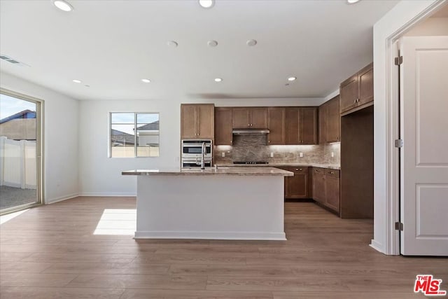 kitchen featuring hardwood / wood-style floors, double oven, black stovetop, an island with sink, and light stone countertops