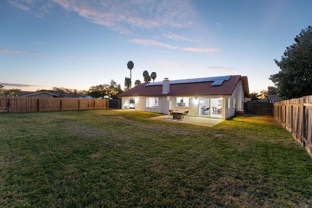 back house at dusk featuring a patio area, a lawn, and solar panels