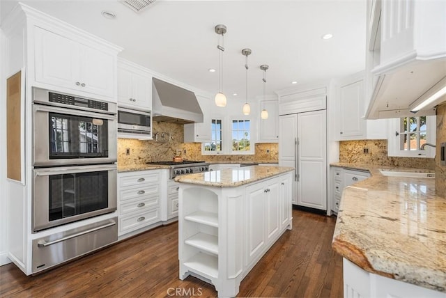 kitchen with wall chimney range hood, a kitchen island, built in appliances, white cabinetry, and hanging light fixtures