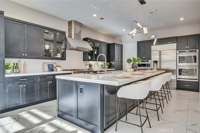 kitchen with sink, tasteful backsplash, appliances with stainless steel finishes, a kitchen island with sink, and wall chimney range hood