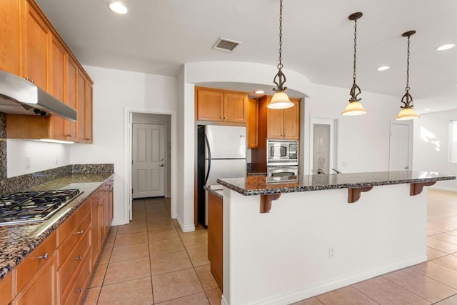 kitchen with appliances with stainless steel finishes, a kitchen bar, hanging light fixtures, and dark stone countertops