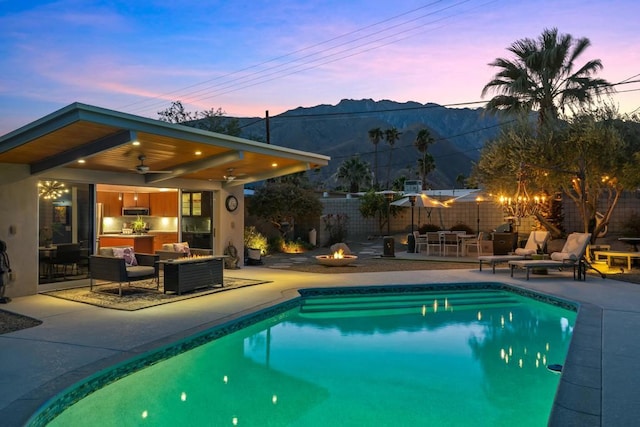 pool at dusk with a mountain view, a patio area, ceiling fan, and an outdoor living space with a fire pit