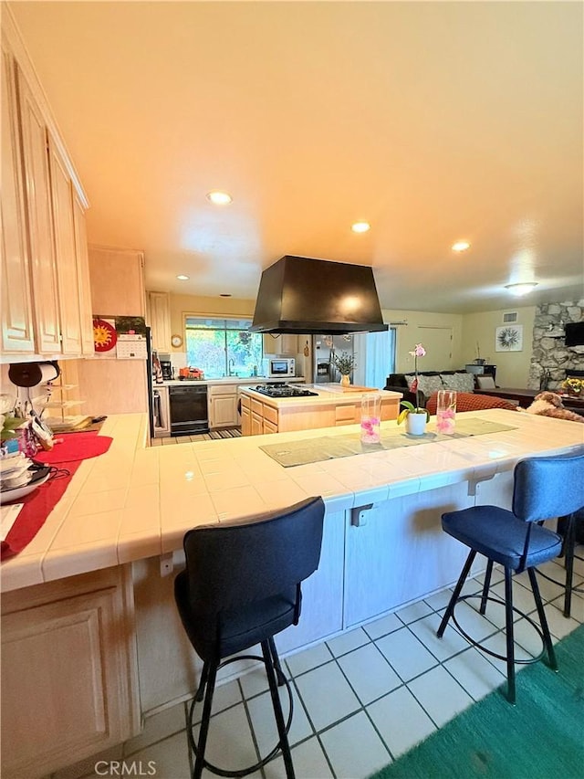 kitchen featuring tile countertops, a kitchen bar, kitchen peninsula, island exhaust hood, and light brown cabinetry