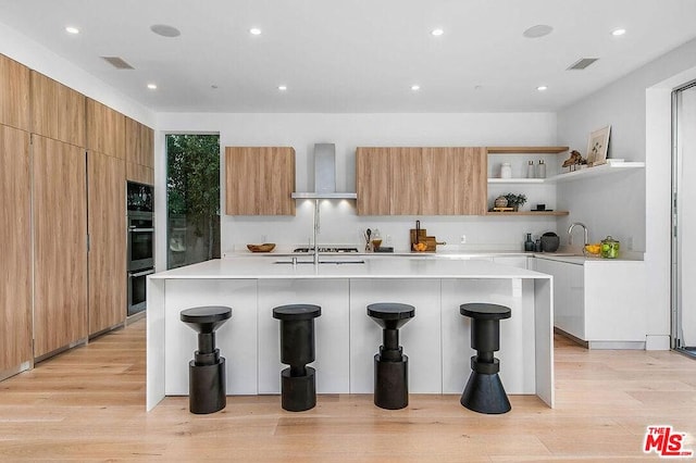 kitchen with wall chimney exhaust hood, a kitchen bar, white cabinetry, light hardwood / wood-style floors, and a kitchen island with sink